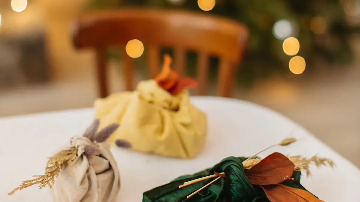 Gifts wrapped in green and beige fabric with dried leaves, furoshiki style, on a table near a Christmas tree